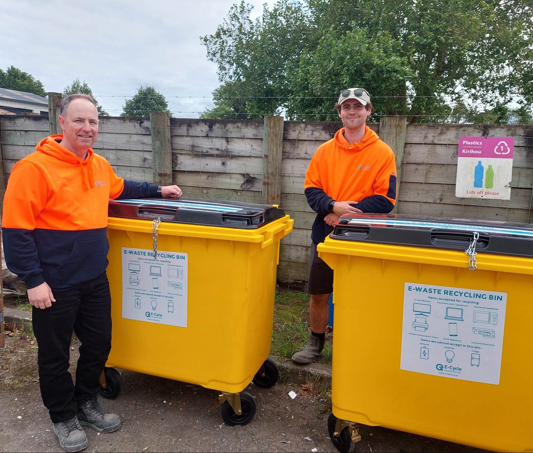 Two workers in high-visibility orange safety gear standing with yellow e-waste recycling bins outdoors. Industrial waste containers feature instructional labels showing acceptable electronic items. Concrete fence and recycling signage visible in background.
