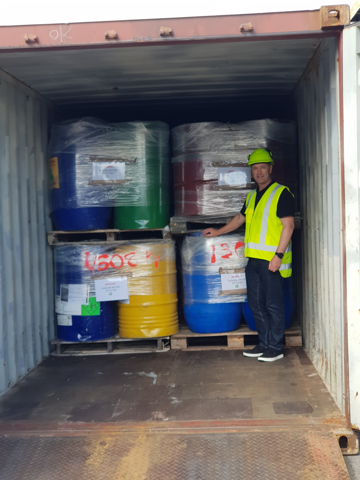 Industrial shipping container loaded with colourful recycling drums on pallets, with worker wearing safety gear including high-visibility vest and hard hat, demonstrating proper handling of electronic waste materials