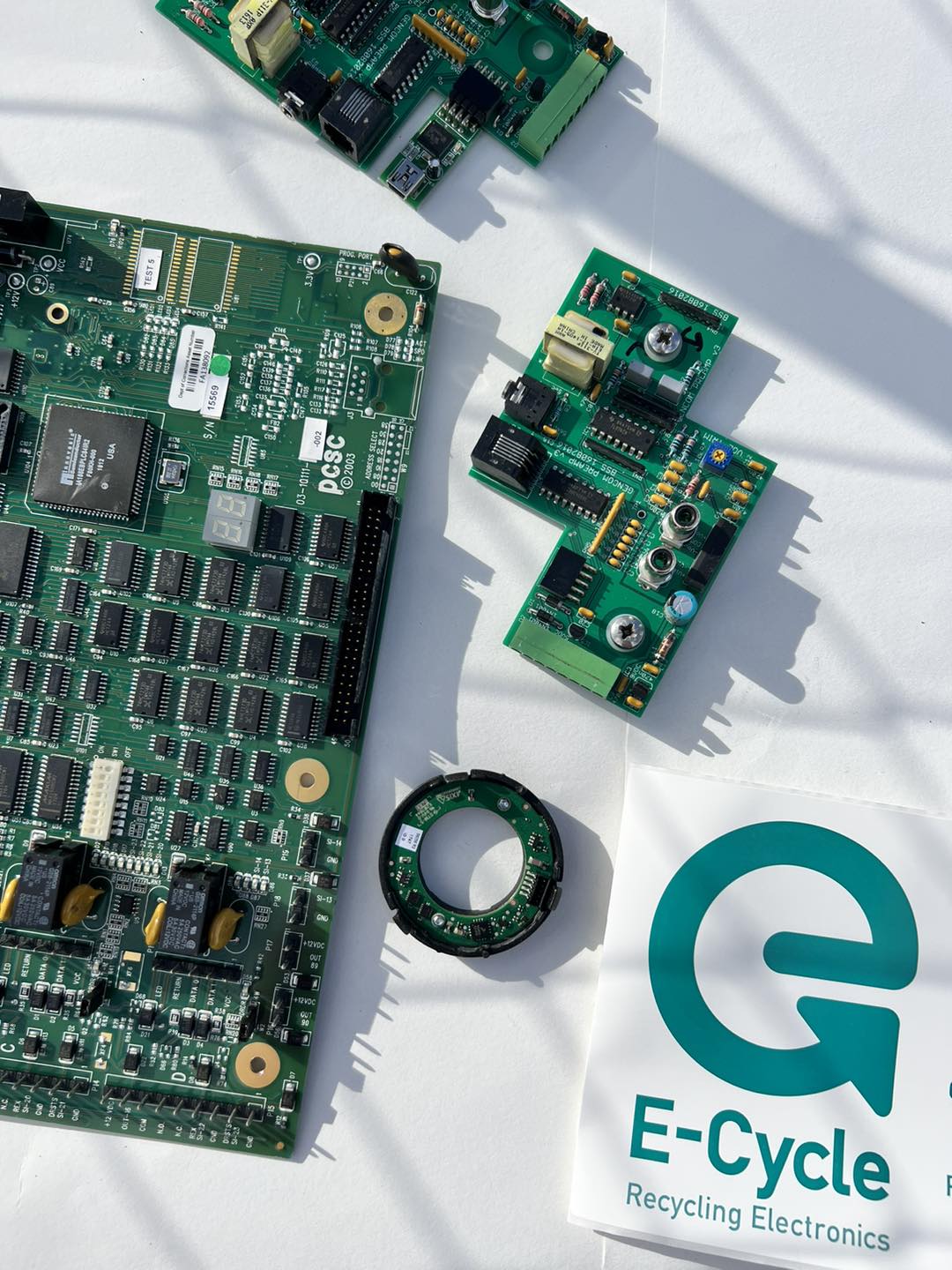 Disassembled electronic circuit boards and components laid out on white surface next to E-Cycle company logo, showcasing electronic recycling process and sustainable e-waste management