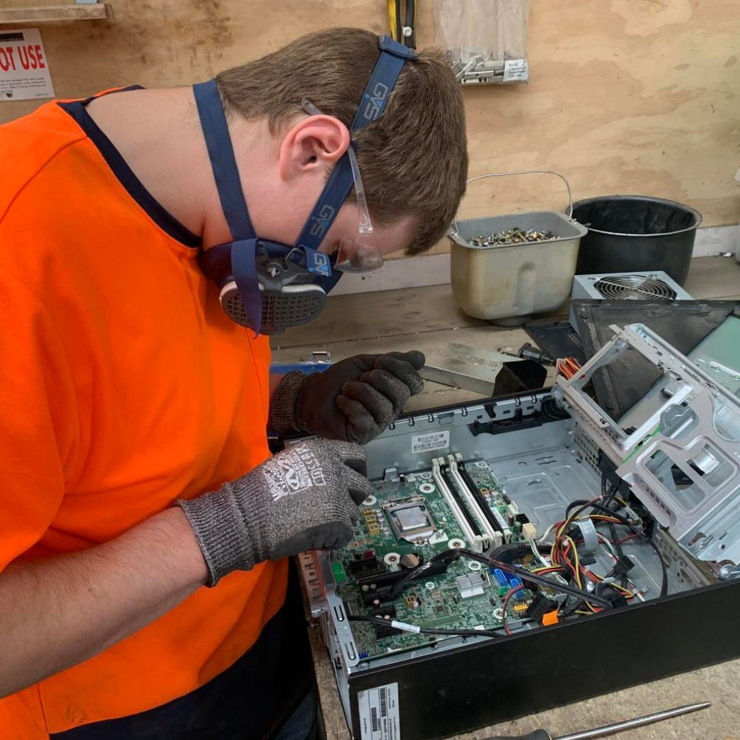 Technician wearing safety gear including respirator mask, orange high-visibility vest, and protective gloves while disassembling electronic equipment and inspecting computer motherboard for recycling process