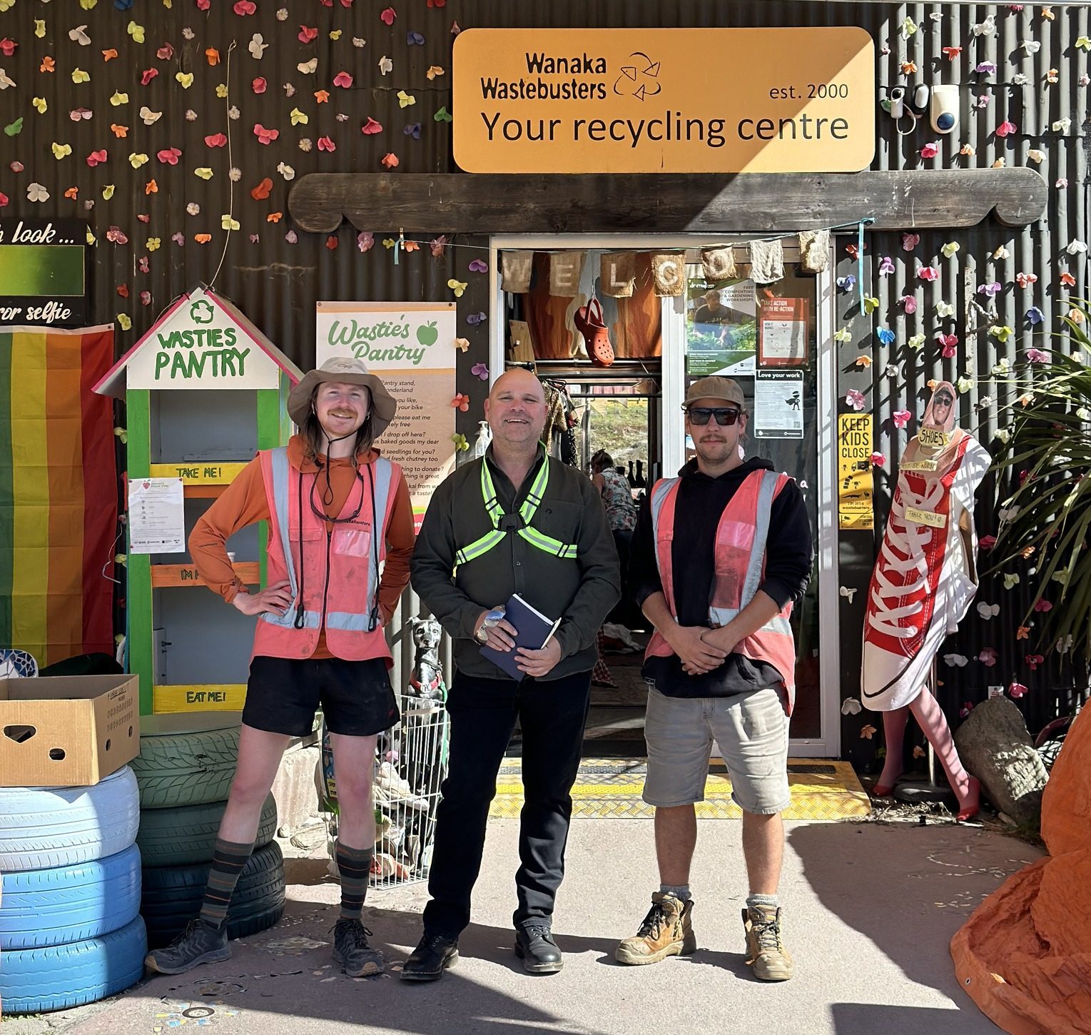 Three workers standing at entrance of local recycling centre with 'Your recycling centre' signage above. Location features colourful decorative elements, community pantry box, and painted tires. Workers wearing safety vests at recycling facility entrance.