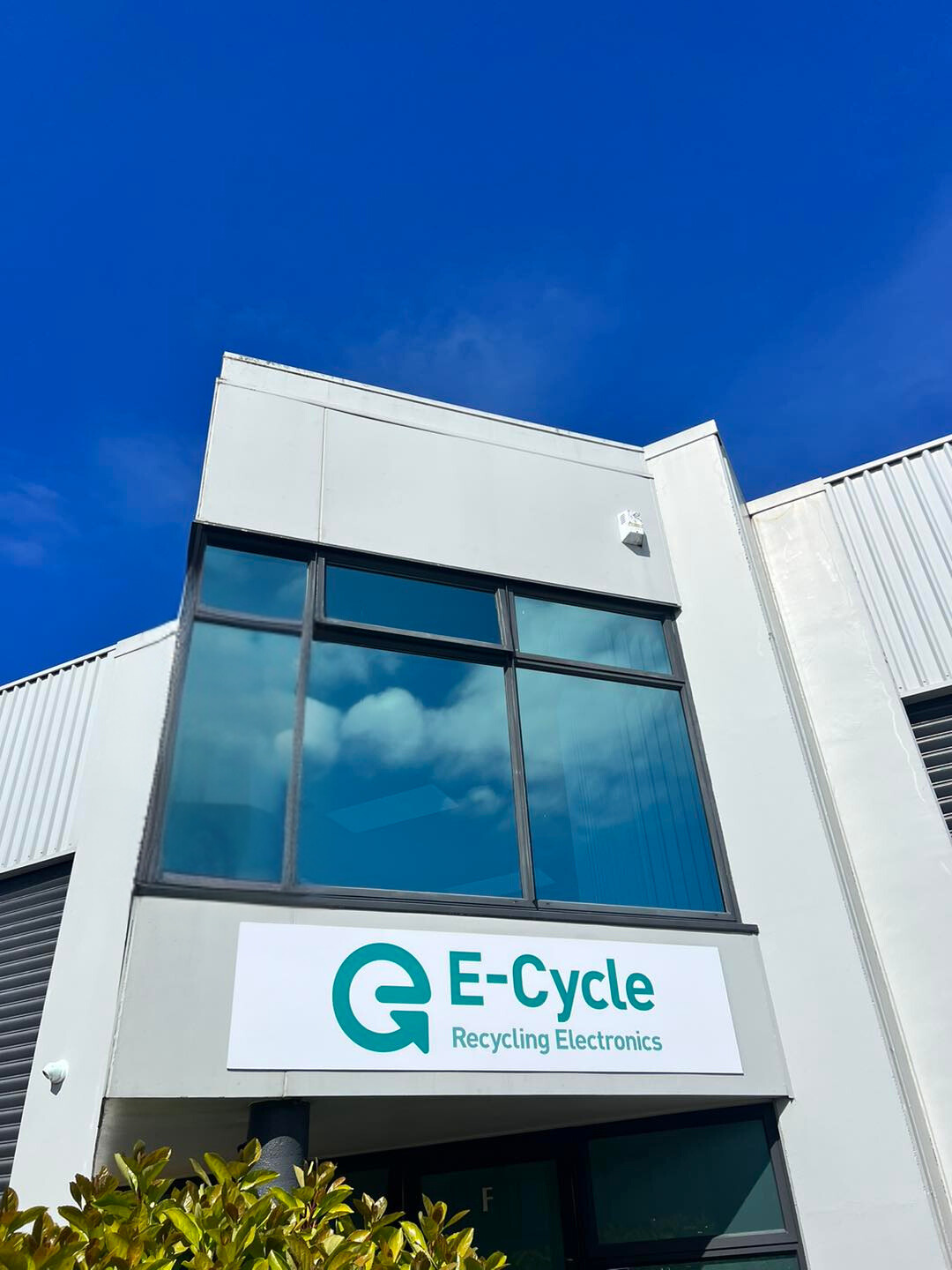 E-Cycle electronics recycling facility exterior with modern white building facade, large reflective windows, and teal company logo signage against vibrant blue sky