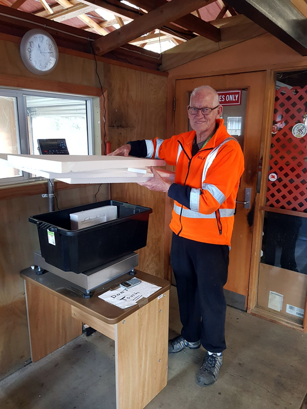 Worker in orange high-visibility safety vest weighing polystyrene on an industrial scale inside a wooden office space. The interior shows a wall clock, wooden panels, and a storage container on a desk, with natural light coming through a skylight.