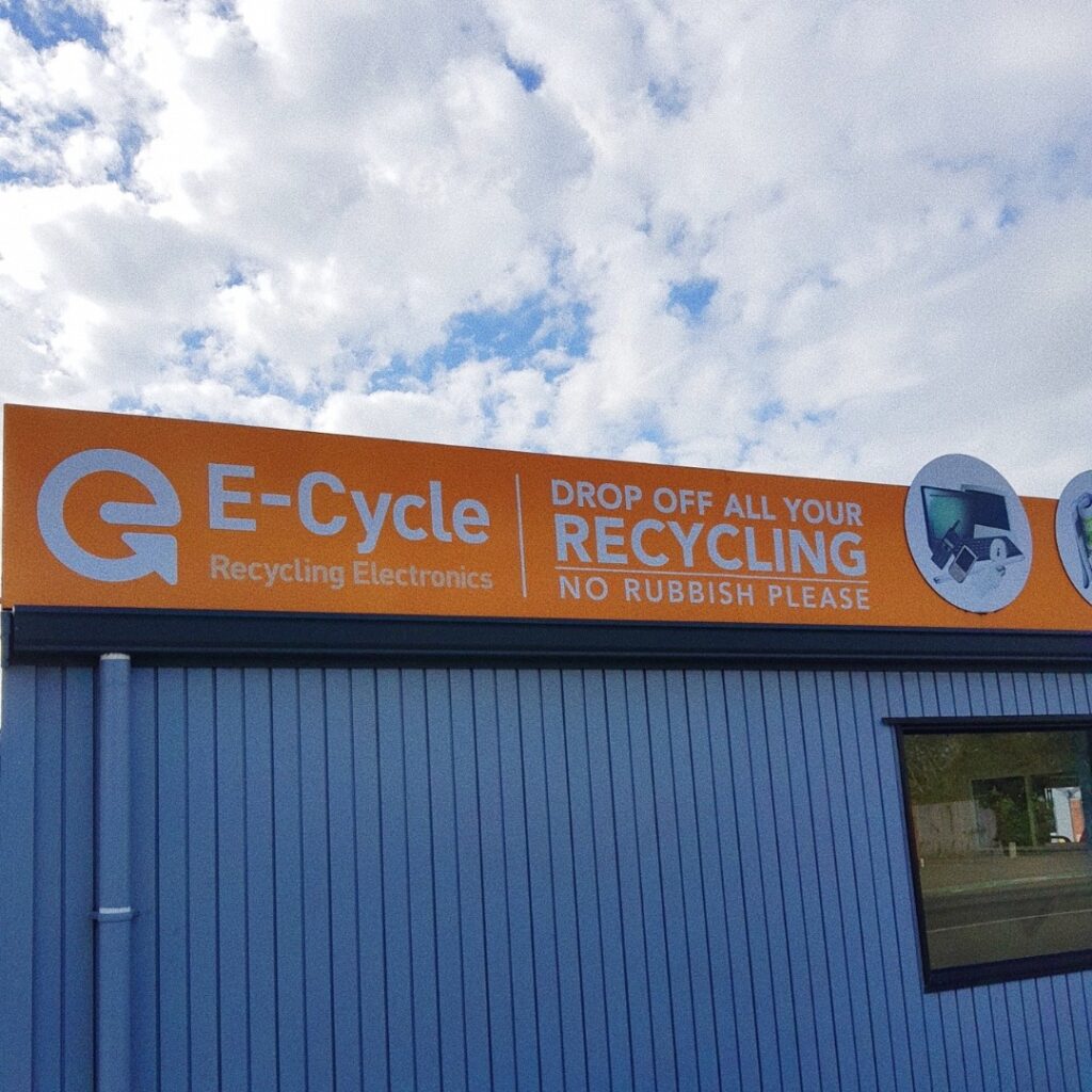 Exterior signage of E-Cycle electronic recycling facility featuring orange branding banner with 'Drop off all your recycling - No rubbish please' message. Blue metal building exterior under cloudy sky. Professional electronics recycling drop-off location.