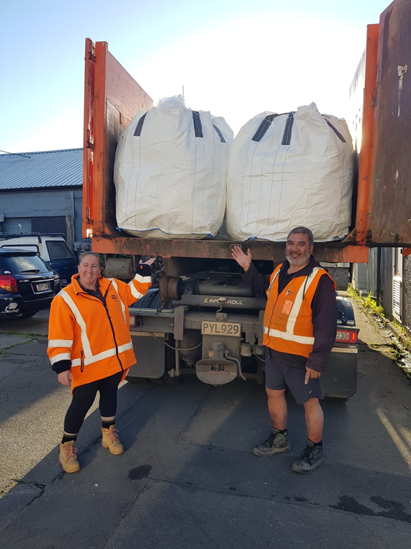 Two workers in high-visibility orange safety gear standing beside a transport truck loaded with large white bulk bags ready for recycling collection