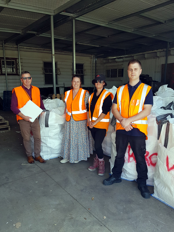 Four warehouse workers wearing high-visibility orange safety vests standing in front of stacked white bulk bags in a recycling facility. The workers are positioned in a line against a concrete wall with overhead lighting.