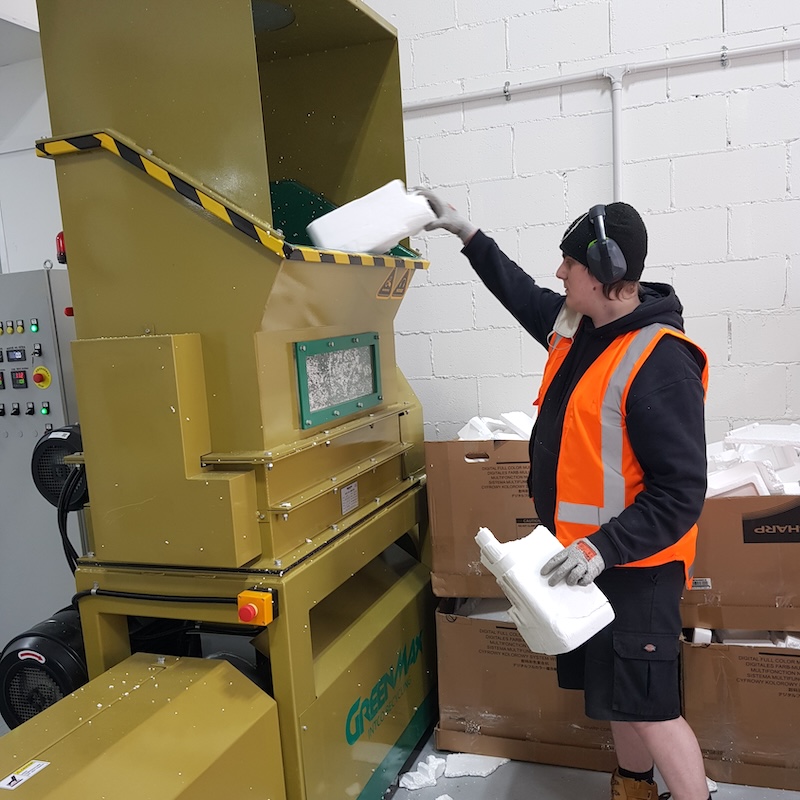 A worker in a black hoodie, orange safety vest, and protective headphones feeds blocks of polystyrene into a large yellow industrial shredding machine. The machine has black and yellow hazard stripes around its feed opening and a control panel mounted on the wall. Several cardboard boxes are stacked nearby in the white-walled industrial space.