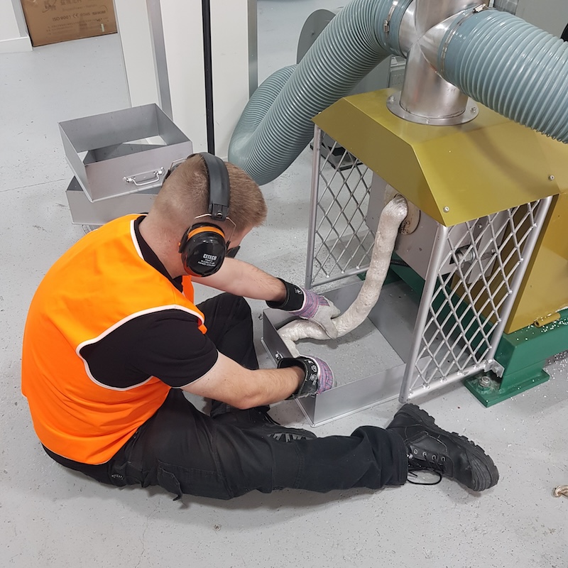 A worker in an orange high-vis shirt and black pants kneels on the ground while servicing industrial equipment. They're wearing protective headphones and working on a machine that has large flexible ducts attached to its top. A metal storage box is visible in the background against a white wall.